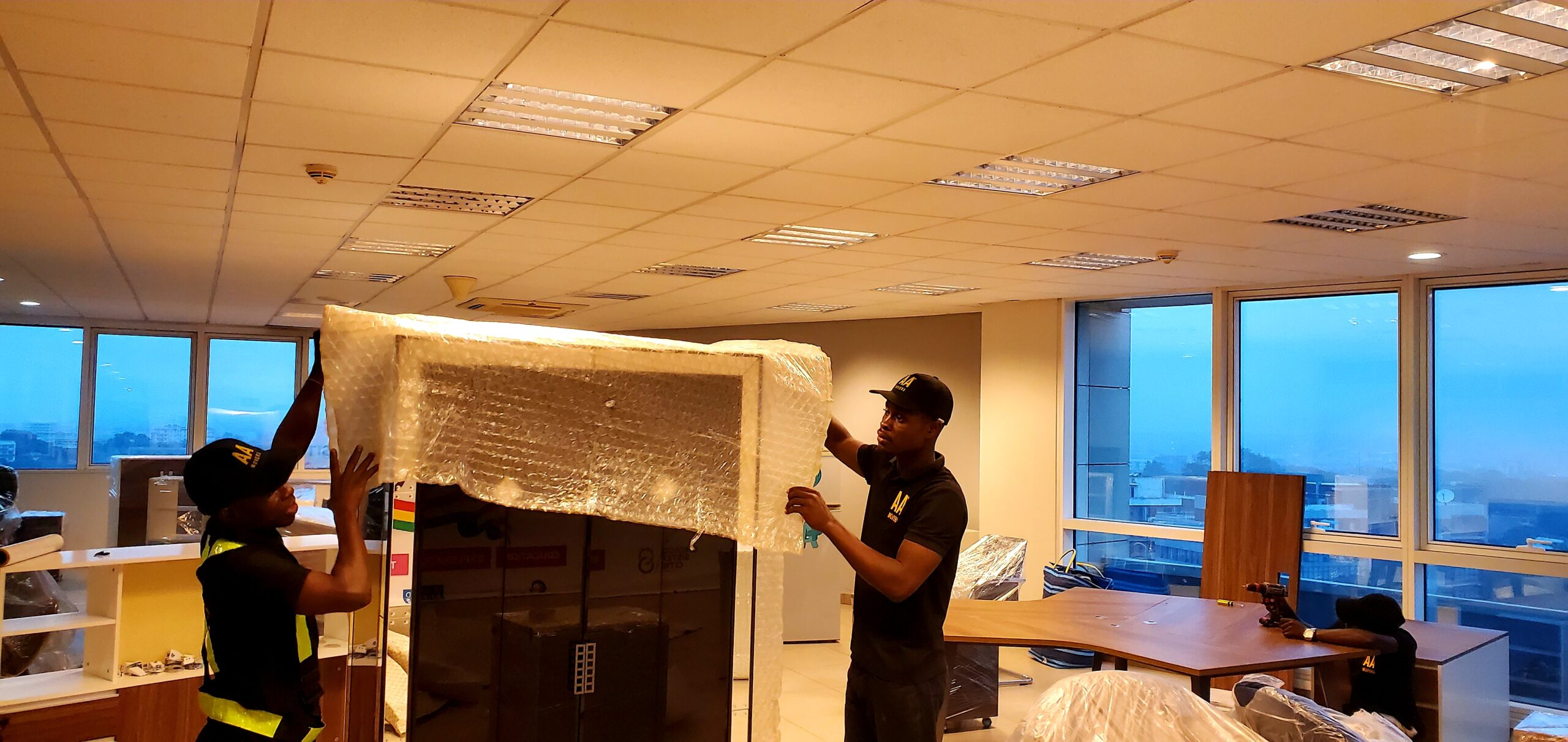 Two neatly uniformed AA+ Movers carefully wrapping a glass and mirror office cabinet with bubble wrap in an office in Ridge, Accra, demonstrating our professional moving services in Ghana for office relocations.
