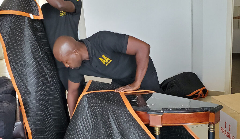 Two AA+ Movers employees in uniform wrapping a marble top side table with a moving blanket, next to a sturdy wooden crate marked 'fragile' with directional arrows, during preparation for an international move.