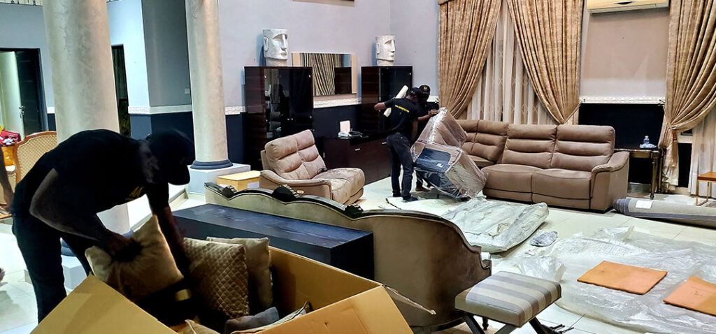 Three AA+ Movers in uniform efficiently packing and wrapping household items during a move, focusing on cushions and a sofa set