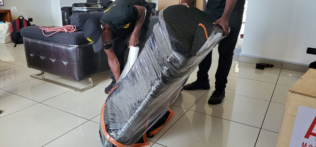 Two AA+ Movers in uniform securing a piece of furniture with a moving blanket and shrink wrap during a relocation